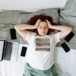 A Stressed Woman Lying on a Bed beside Cellphones and a Laptop