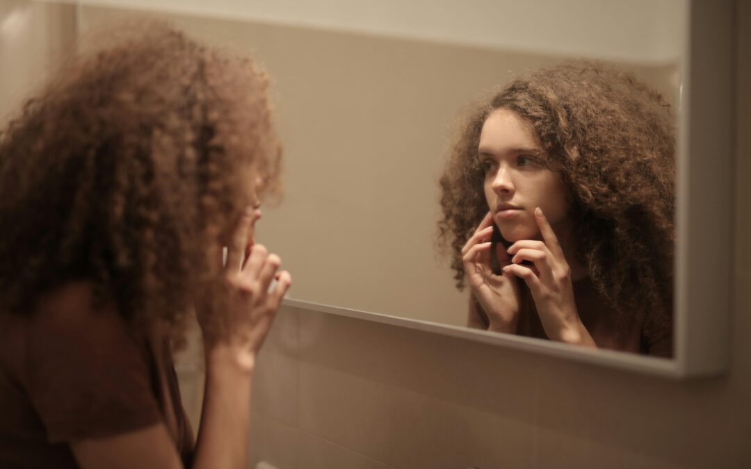 Serious young lady touching face while looking at acne in mirror in bathroom