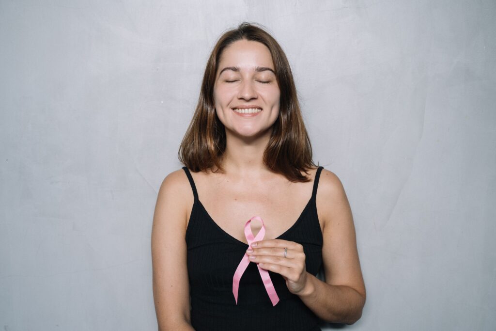 A smiling woman with eyes closed holds a pink breast cancer awareness ribbon, symbolizing hope and health