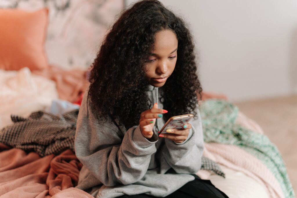 A teenage girl with curly hair using a smartphone while sitting on a bed indoors