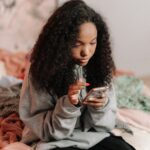A teenage girl with curly hair using a smartphone while sitting on a bed indoors